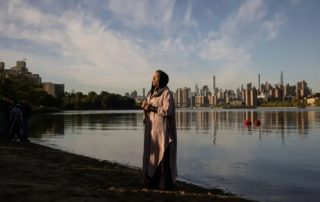 Woman stands along river bank with cityscape in the background